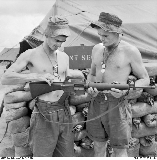 Bien Hoa, Vietnam. 1965-09. Two bare-chested Australians Corporal Lex McAulay (left) of Innisfail, Qld, and Corporal John Henderson of Macquarie Fields, NSW, inspect an Armalite rifle at the headquarters of the 1st Battalion, The Royal Australian Regiment (1RAR). Photograph: Bryan Dunne. Australian War Memorial Accession Number DNE/65/0335A/VN
