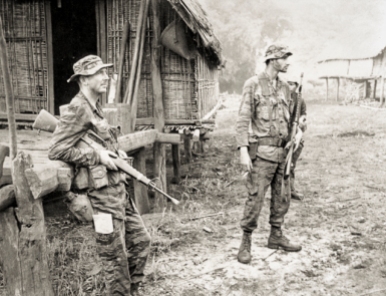 Pleiku, South Vietnam. July 1970. Australian Army Training Team Vietnam (AATTV) adviser, Captain (Capt) Peter Shilston of Williamtown, NSW (left), and an American adviser look on while Montagnard soldiers of the 1st Battalion, 2nd Mobile Strike Force search a village during an operation in central South Vietnam. Capt Shilston is commander of the Battalion which operates out of Pleiku. Photo: John Fairley. Australian War Memorial Accession Number: P00963.063